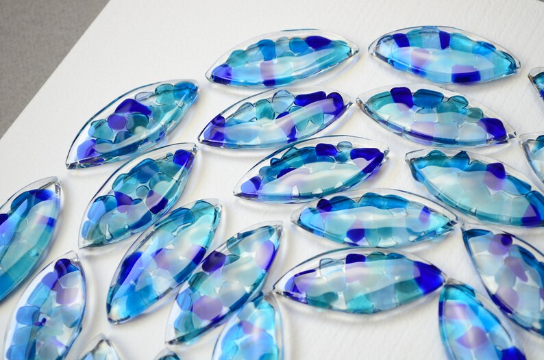 May include: A collection of blue and purple glass pieces shaped like teardrops, each filled with smaller pieces of blue and purple glass. The pieces are arranged in a circular pattern on a white surface.