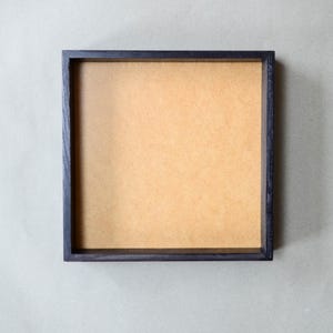May include: A square shadow box frame featuring a dark, stained wooden border and a flat, light brown fiberboard backing. The frame has a clean, geometric design with straight edges and a deep interior space for displaying various objects.
