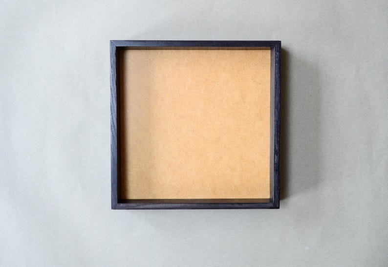 May include: A square shadow box frame featuring a dark, stained wooden border and a flat, light brown fiberboard backing. The frame has a clean, geometric design with straight edges and a deep interior space for displaying various objects.
