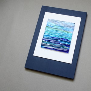 May include: A framed abstract art piece with a blue, green, and white glass mosaic design. The frame is dark blue and the mat is white.