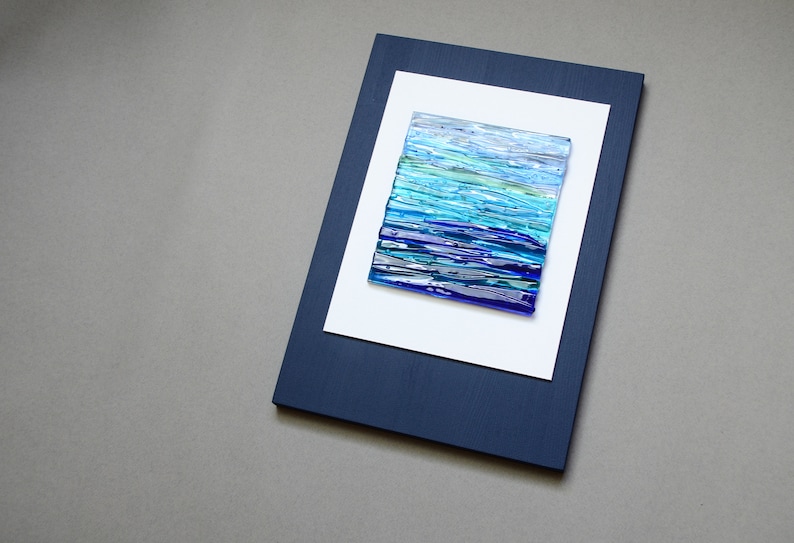 May include: A framed abstract art piece with a blue, green, and white glass mosaic design. The frame is dark blue and the mat is white.
