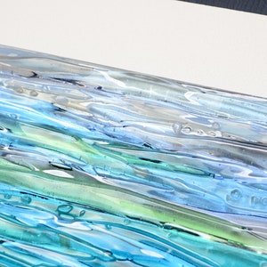 May include: A rectangular glass tile with a layered design of blue, green, and white glass. The glass has a wavy, textured surface with air bubbles trapped inside.