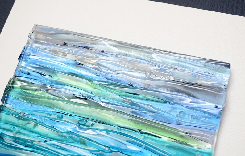 May include: A rectangular glass tile with a layered design of blue, green, and white glass. The glass has a wavy, textured surface with air bubbles trapped inside.