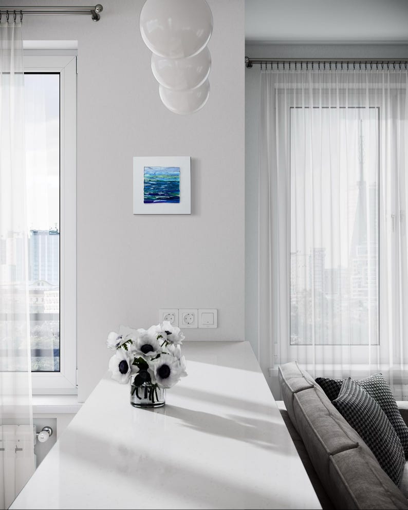 May include: A modern interior featuring a white countertop with a vase of white flowers with black centers. A small square painting with blue and green abstract art hangs on the wall. A gray sofa and sheer curtains are visible.