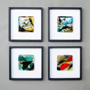 May include: Four square wall art pieces in black frames, each containing a unique fused glass tile. The tiles feature abstract patterns in various colors including yellow, white, black, blue, teal, red, and brown, set against a white background.