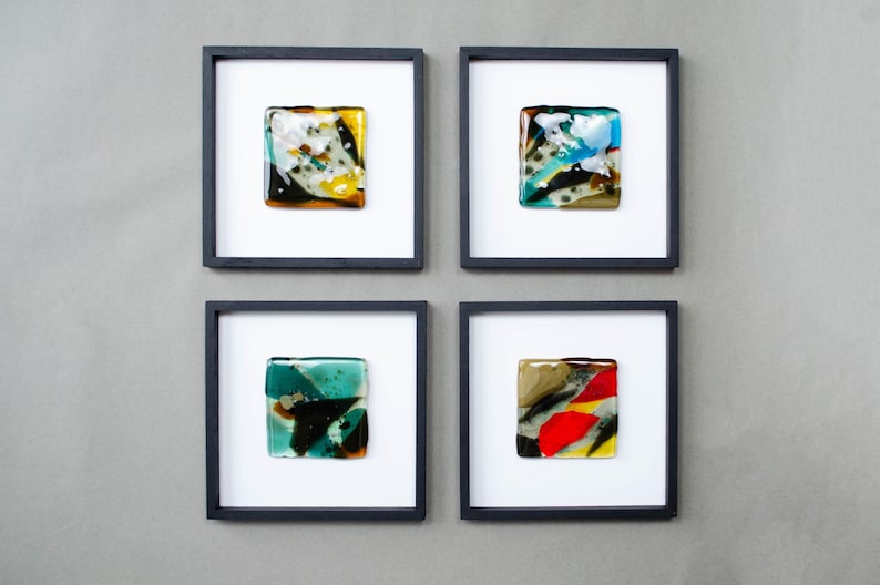 May include: Four square wall art pieces in black frames, each containing a unique fused glass tile. The tiles feature abstract patterns in various colors including yellow, white, black, blue, teal, red, and brown, set against a white background.
