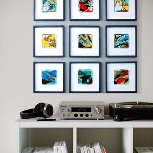May include: A grid of nine square wall art pieces featuring colorful fused glass tiles in dark blue frames, displayed above a white media console with a silver stereo receiver, black headphones, a turntable, and stacks of books on shelves.