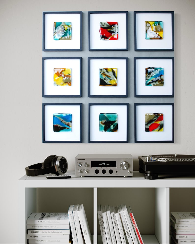 May include: A grid of nine square wall art pieces featuring colorful fused glass tiles in dark blue frames, displayed above a white media console with a silver stereo receiver, black headphones, a turntable, and stacks of books on shelves.