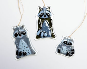 May include: Three glass ornaments shaped like raccoons. The ornaments are blue, black, and green. They are hanging from twine.