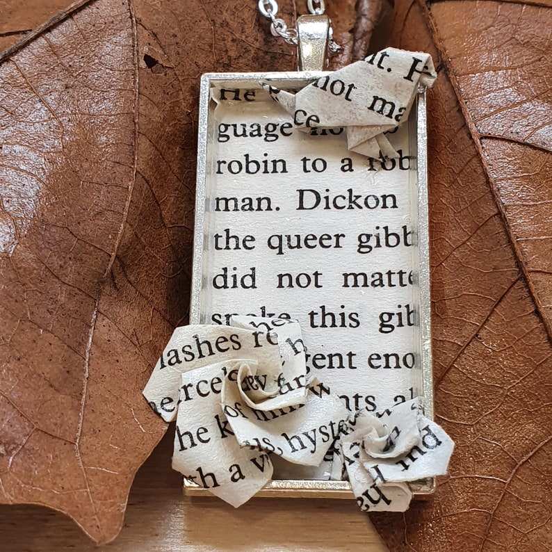 Op de afbeelding: Een zilveren ketting met een rechthoekige hanger met een stuk papier met tekst. Het papier is gevouwen tot een bloemvorm en is gedeeltelijk zichtbaar buiten het frame. De hanger ligt op een bruine ondergrond met een bruin blad.