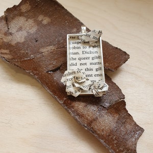 Op de afbeelding: Een zilveren hanger met een rechthoekig frame met een gedrukt tekste excerpt. Het frame is versierd met twee papieren bloemen gemaakt van hetzelfde bedrukte papier.