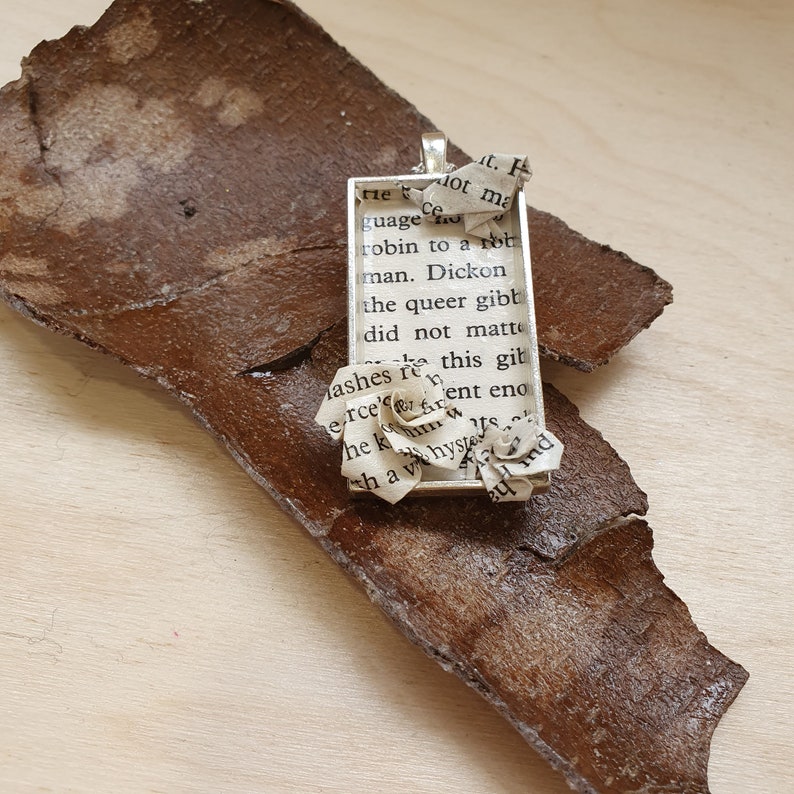 Op de afbeelding: Een zilveren hanger met een rechthoekig frame met een gedrukt tekste excerpt. Het frame is versierd met twee papieren bloemen gemaakt van hetzelfde bedrukte papier.