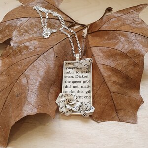 Op de afbeelding: Een zilveren ketting met een rechthoekige hanger met een papieren bloem en tekst uit een boek. De tekst luidt "lot ma guage robin to a tot man. Dickon the queer gibt did not matte st ashes re n erce S he ko this gibt ent eno ats shyste hav  md".