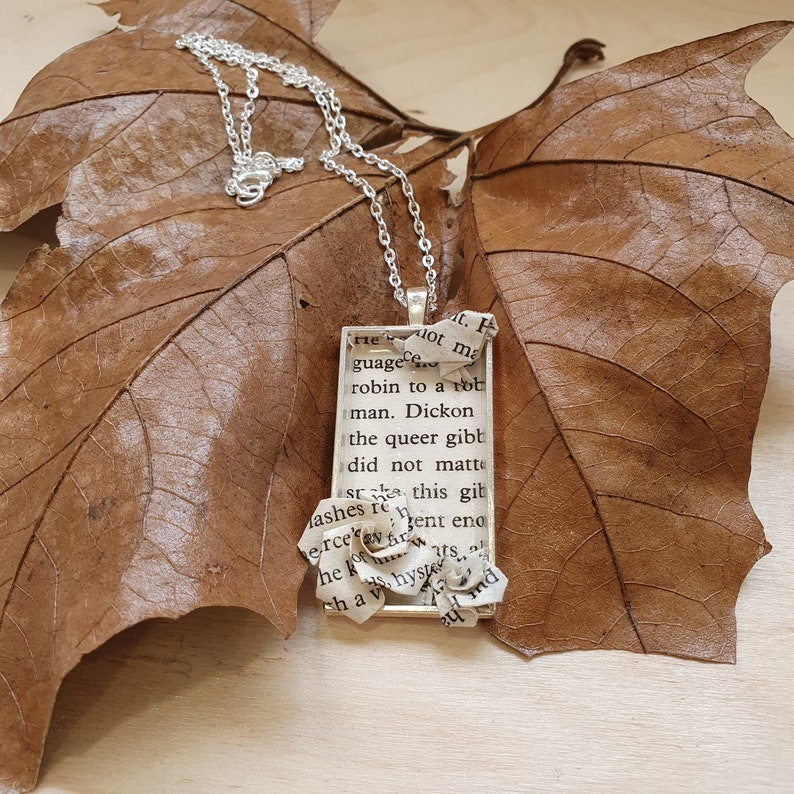 Op de afbeelding: Een zilveren ketting met een rechthoekige hanger met een papieren bloem en tekst uit een boek. De tekst luidt "lot ma guage robin to a tot man. Dickon the queer gibt did not matte st ashes re n erce S he ko this gibt ent eno ats shyste hav  md".