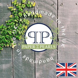 Op de afbeelding: Een witte cirkel met de letters "QP" in blauw en de tekst "PAPER & PEARLS" in zwart. De cirkel is omgeven door de tekst "Handmade in the UK" in wit op een grijze houten achtergrond met groene wijnstokken.