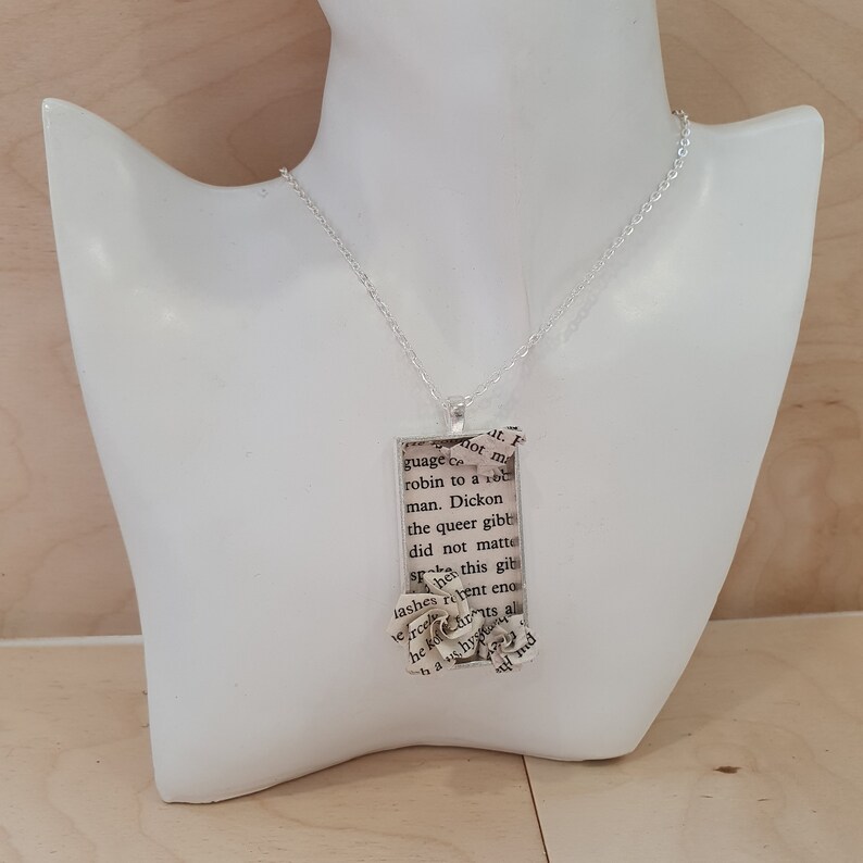 Op de afbeelding: Een zilveren ketting met een rechthoekige hanger met een boekpagina met de tekst "not m guage robin to a o man. Dickon the queer gibb did not matte this gib SEA lashes rent eng verode he khits al h hy md", de tekst is gedeeltelijk bedekt door een papieren bloem.