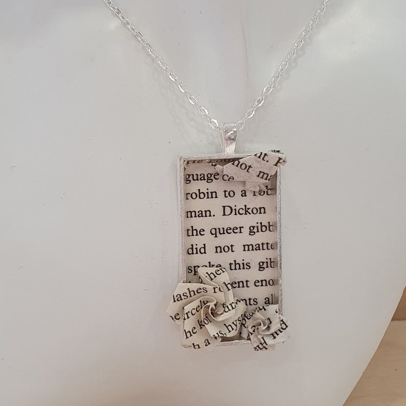 Op de afbeelding: Een zilveren ketting met een rechthoekige hanger met een papieren bloem en tekst uit een boek. De tekst luidt "at. F mot ma guage ce robin to a 100 man. Dickon the queer gibt did not matte sp then this git lashes rebent eno ercel he kofarts al haus, hyst d"