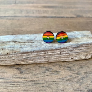 以下が含まれることがあります： 虹色の木製のイヤリングのペア。各イヤリングの中央には小さなハートがあります。イヤリングは明るい色の木製の表面の上にあります。