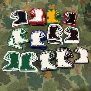 May include: A collection of embroidered boot patches in various colors, including black, white, red, yellow, green, and blue. Each patch features a detailed boot design with a tassel, outlined in a contrasting color. The patches are arranged on a camouflage background.
