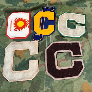 Peut inclure: Plusieurs écussons de veste de lettres vintage. Les écussons en forme de lettre "C" sont de différentes couleurs : blanc, jaune, vert et marron. Un écusson porte le mot "ORCHESTRA" et un motif de soleil.
