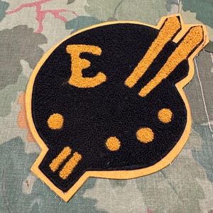 May include: A black patch with a yellow border and accents. The patch features the letter "E" in yellow, along with two parallel lines and three dots. The patch is on a camouflage background.
