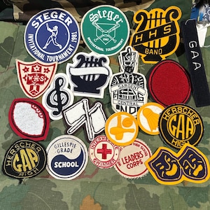 Puede incluir: Una colección de varios parches de tela bordados con diversas formas y diseños, incluyendo logotipos deportivos, instrumentos musicales, emblemas escolares y texto como Steger Invitational Tournament, HHS Band, GAA, WHS y Herscher Gap High.