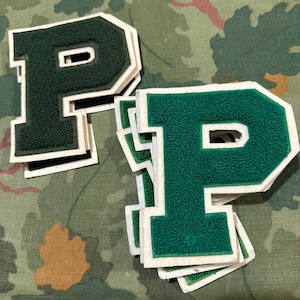 Puede incluir: Varios parches de fieltro verde con la letra "P" y bordes blancos están apilados sobre un fondo de camuflaje. Los parches tienen un estilo clásico de letra universitaria, con una apariencia texturizada y difusa. La letra "P" es un diseño audaz y en bloque.
