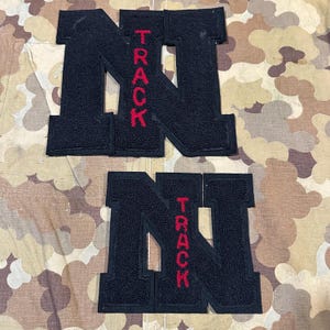 Op de afbeelding: Twee zwarte vilt patches in de vorm van de letter "N" met het woord "TRACK" in rode letters. De patches liggen op een camouflage achtergrond.