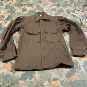 May include: An olive green wool shirt with two chest pockets and button fastenings. The long-sleeved shirt has a collared neckline and buttoned cuffs. A measuring tape is visible in the background. The shirt is laid flat on camouflage fabric.