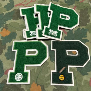 Puede incluir: Parches de letras universitarias verdes y blancas con forma de letra "P". Algunos parches tienen iconos de temática deportiva, como una pelota de béisbol, un bate y una pelota de béisbol, y un emblema de espadas cruzadas. Los parches están sobre un fondo de camuflaje.