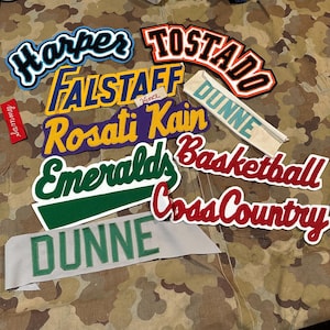 Może przedstawiać: Kolekcja haftowanych naszywek w różnych kolorach i czcionkach. Naszywki zawierają imiona takie jak "Harper", "Falstaff" i "Rosati Kain", a także terminy sportowe, takie jak "Basketball" i "Cross Country". Tło to wzór kamuflażu.