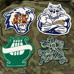May include: Four embroidered patches featuring different designs. The first patch is a white panther with a blue outline and a red "LAKE" and "M" in the center. The second patch is a blue wolf with a white outline and sunglasses, holding a football. The third patch is a green lyre with a white outline. The fourth patch is a green knight riding a horse with a white outline.