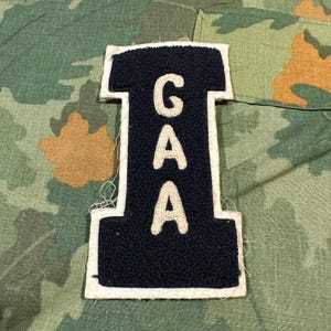 May include: A navy blue varsity letter patch in the shape of the letter I, featuring the white embroidered letters G, A, and A stacked vertically in the center. The patch has a thick white border and a textured, fuzzy chenille-style surface.