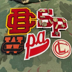 May include: A collection of five fabric patches including a large red and yellow BC monogram, a red and white SP patch with gold metallic pins, an orange and black WC monogram, a red and white script pg patch, and a circular white and red CL patch.