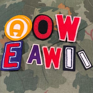 May include: A collection of felt fabric patches featuring various letters and shapes. The set includes a yellow and white circular patch with an A, a black circular patch, a large red W, a blue and red E, a purple A, a red W, and two rectangular patches.