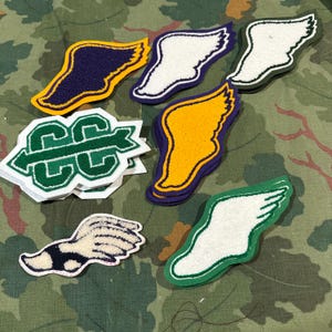 Puede incluir: Surtido de parches bordados, incluyendo alas y un logo "CC". Los parches de alas presentan diseños en blanco, amarillo y azul marino con bordes contrastantes. El parche "CC" es verde y blanco.