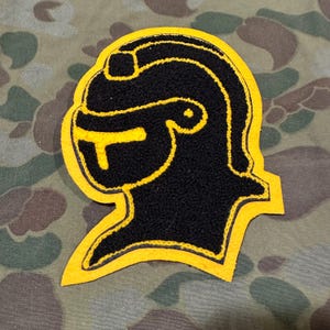 May include: A black and yellow felt patch depicting a knight's helmet in profile.