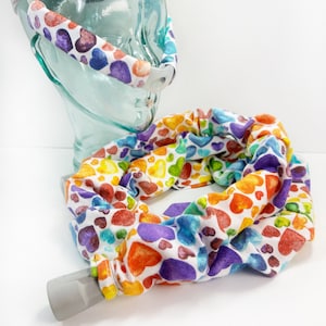 May include: A white fabric tube with a rainbow heart print. The tube is designed to cover a CPAP hose. The tube is shown wrapped around a clear glass CPAP mask.