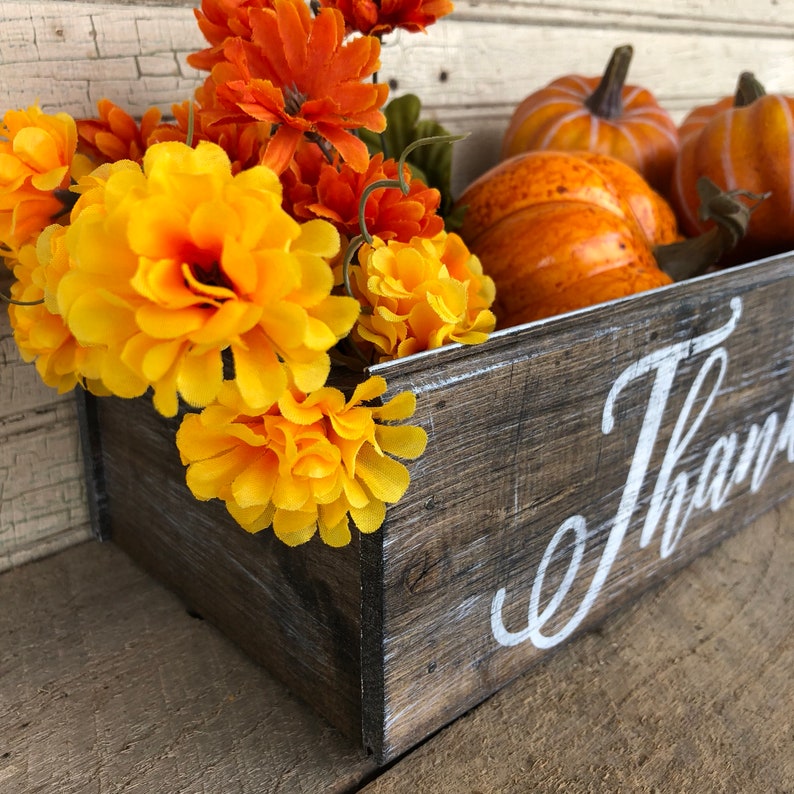 May include: A wooden box with the word "Thank" painted in white on the side. The box is filled with orange and yellow flowers and small orange pumpkins.