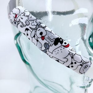 May include: A white fabric eyeglass strap featuring a repeating pattern of cartoon dogs in various poses. The dogs are primarily white with black spots and accents, and some have red tongues. The strap is attached to a clear, glass object.