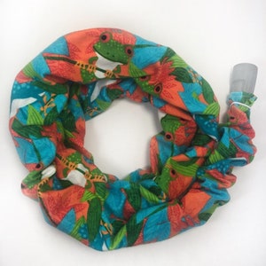 May include: A colorful fabric neck gaiter featuring a repeating pattern of green frogs, blue leaves, and red flowers on an orange background. The gaiter has a clear plastic tube attached to one end.