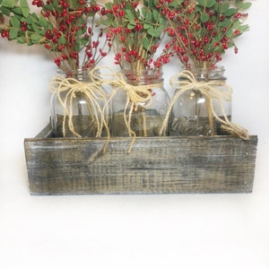 Puede incluir: Tres tarros de vidrio transparentes con ramitas de bayas rojas y lazos de cuerda se muestran en una caja de madera r&uacute;stica.