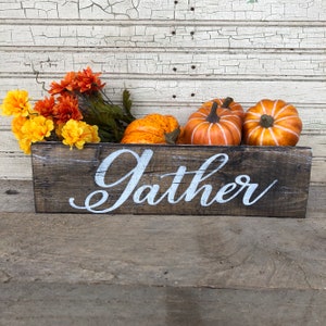 May include: A rustic wooden box with the word "Gather" painted in white. The box is filled with orange and yellow fall-themed flowers and small orange pumpkins.
