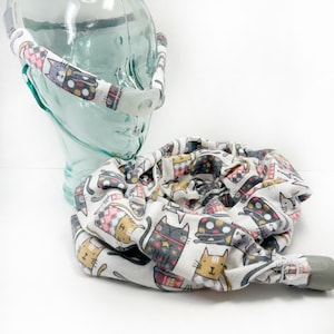 May include: A white fabric CPAP mask with a cat print. The mask has a clear plastic tube and a gray plastic connector.