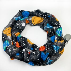 May include: A black and white fabric neck gaiter featuring a colorful space-themed design. The design includes planets, stars, and the sun. The gaiter has a gray plastic clasp.