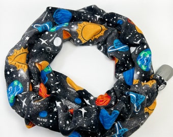 May include: A black and white fabric neck gaiter featuring a colorful space-themed design. The design includes planets, stars, and the sun. The gaiter has a gray plastic clasp.