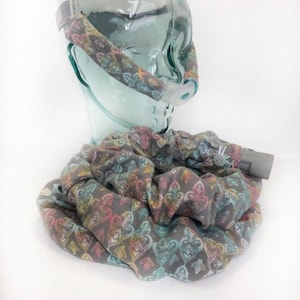 May include: A gray CPAP mask cover and tubing wrap with a colorful floral pattern. The fabric is adorned with pink, yellow, and teal accents. A button secures the wrap. The mask is on a clear glass head form.