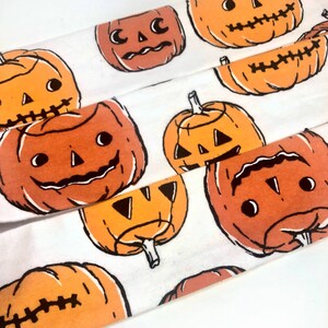 May include: White fabric with a repeating pattern of orange jack-o'-lantern faces with black outlines. The pumpkins have various expressions and are in different sizes and orientations, perfect for Halloween.