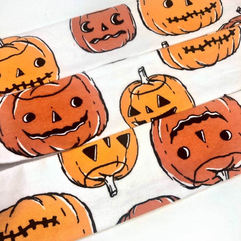 May include: White fabric with a repeating pattern of orange jack-o'-lantern faces with black outlines. The pumpkins have various expressions and are in different sizes and orientations, perfect for Halloween.