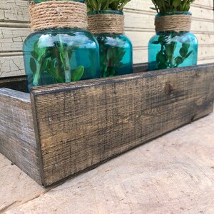 May include: Three teal blue glass jars with greenery inside, sitting in a rustic wooden planter box. The planter box is made of dark stained wood with a distressed finish.